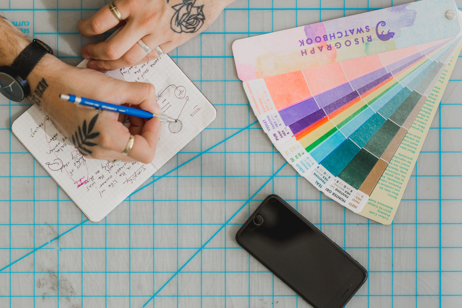 Tattooed hands sketch in a grid notebook beside a Risograph swatchbook and smartphone on a gridded cutting mat, suggesting design planning.