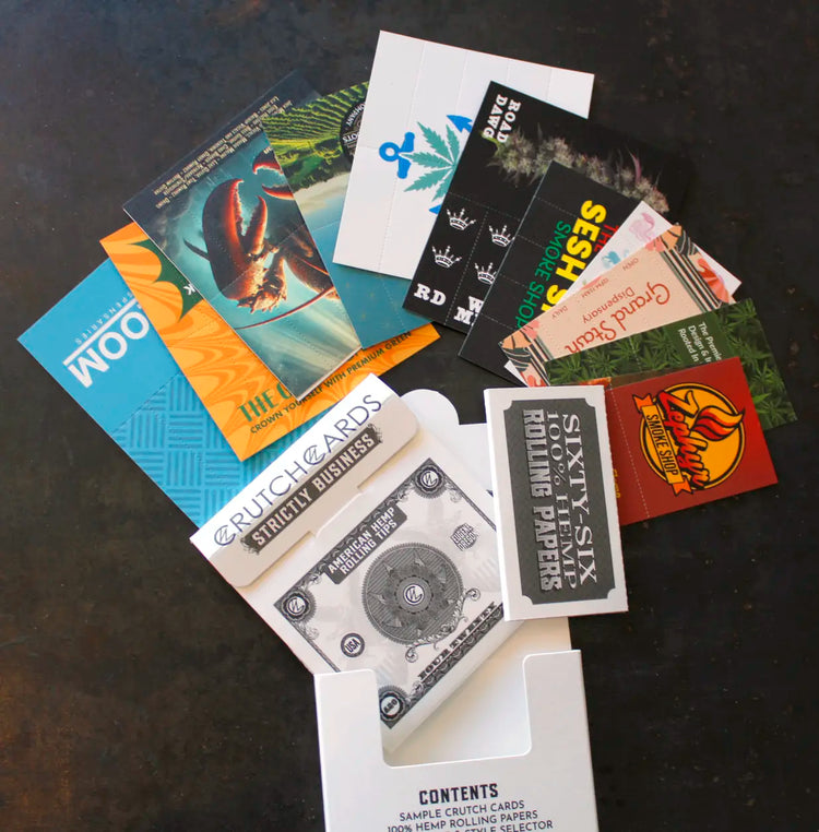 Open Crutch Cards sample kit on a dark surface, displaying a spread of colorful branded Crutch Cards, a pack of “Sixty-Six” hemp rolling papers, and the kit folder labeled “Strictly Business.