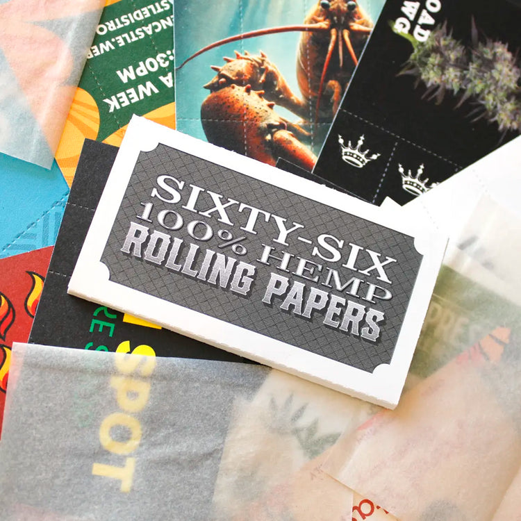 Pack of “Sixty-Six 100% Hemp Rolling Papers” placed on top of assorted Crutch Cards and translucent rolling papers. The background includes bold, colorful designs and cannabis-themed artwork.