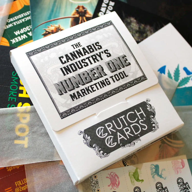 Close-up of a Crutch Cards sample kit box with bold text reading “The Cannabis Industry’s Number One Marketing Tool.” The box sits atop various branded Crutch Cards, rolling papers, and design samples, partially visible in the background.