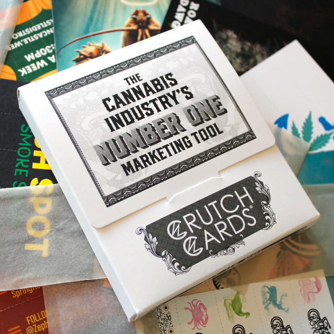Close-up of a Crutch Cards sample kit box with bold text reading “The Cannabis Industry’s Number One Marketing Tool.” The box sits atop various branded Crutch Cards, rolling papers, and design samples, partially visible in the background.