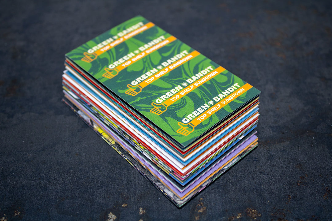 Stack of colorful custom Crutch Cards on a dark tabletop, featuring "Green Bandit Top Shelf Sungrown" designs on top.