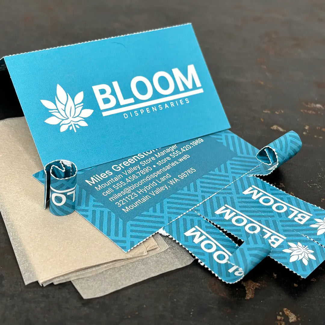 Close-up of a blue Crutch Card for Bloom Dispensaries, partially torn to reveal perforated rolling tips. Some tips are rolled up and standing upright on a black surface, with translucent hemp rolling papers underneath. The card displays business contact information and branding.