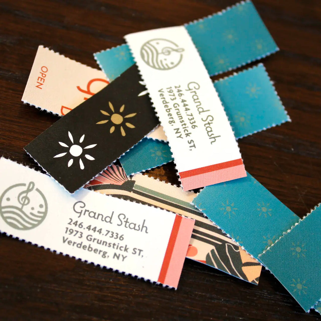 A pile of rolling tips, perforated Crutch Card tips with various designs lies on a dark wood surface, including white tags for "Grand Stash" in Verdeberg, NY, and black strips decorated with minimalist sun icons.