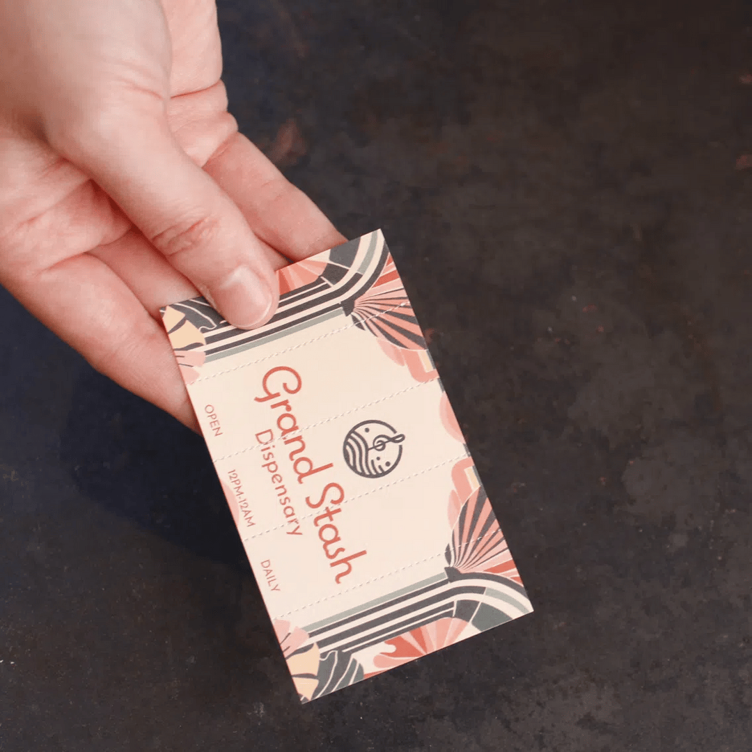 A hand holds and tears a cream-colored Crutch Card for "Grand Stash Dispensary" featuring a retro art deco floral design and perforated edges, indicating it can be torn into individual filter tips.